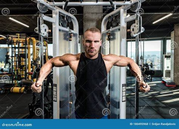 young active strong sweaty fit muscular man with big muscles sitting on the bench and doing biceps workout with dumbbells in the gym as hardcore cross training